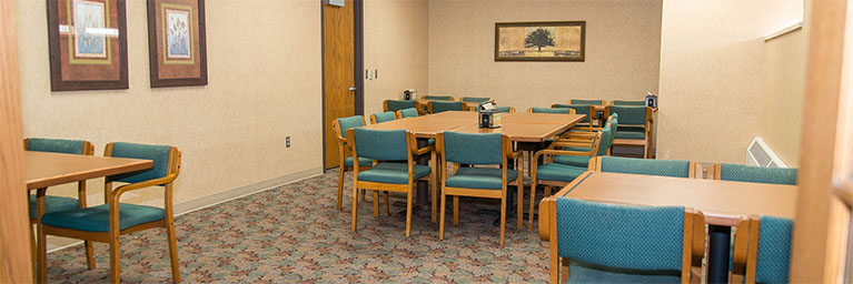 A small meeting or dining room at Bethany Lutheran College featuring tables with green chairs, beige walls, and framed artwork.