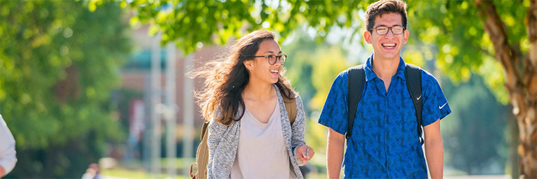 Two smiling students with backpacks walk outdoors on a sunny day, enjoying the vibrant atmosphere of the Minnesota campus.