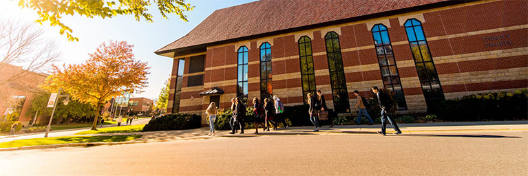 A group walks past a Mankato campus building with tall windows and nearby autumn trees on a sunny day.
