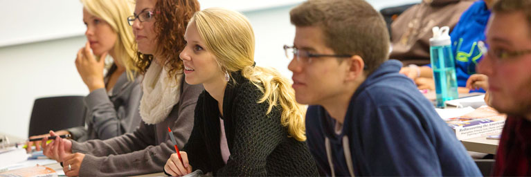A group of students sit at desks in a classroom, attentively listening and taking notes.