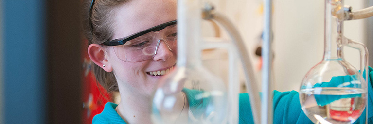 A smiling individual at Bethany Lutheran College uses glass laboratory equipment and clear liquids on campus.