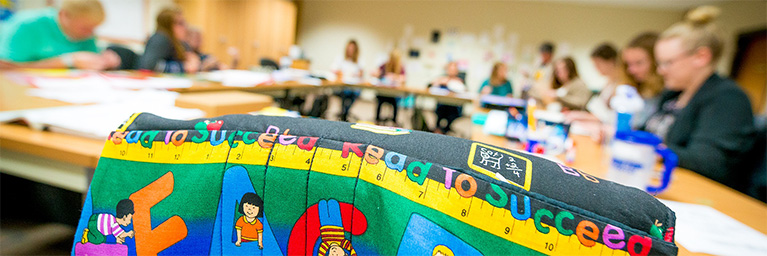 “Colorful Road to Succeed fabric draped over a chair, with a campus community meeting visible in the background.”.