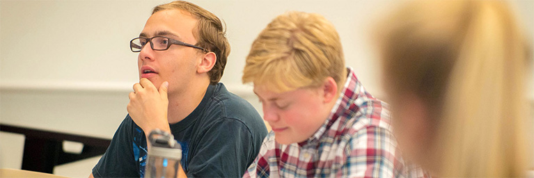 Two students at Bethany Lutheran College in Mankato sit in a classroom; one listens attentively while the other concentrates on his work.