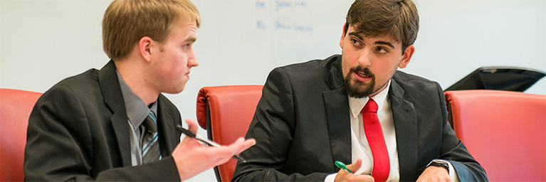 Two men in suits sit at a table, discussing student engagement, with a whiteboard visible in the background.