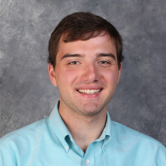 A Bethany Lutheran College student with short brown hair smiles at the camera, wearing a light blue collared shirt against a gray backdrop.
