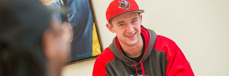 Young man wearing a red and gray hoodie and cap smiles indoors, representing student engagement at Bethany Lutheran College in Minnesota.