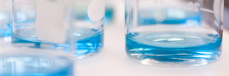 Close-up of clear glass beakers containing blue liquid on a white surface at Bethany Lutheran College in Mankato.