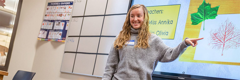 A woman in a gray sweater points to a leaf diagram displayed on a screen in a Minnesota classroom, engaging students.