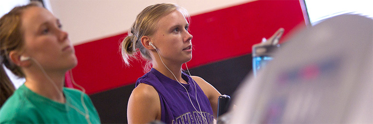 Two women wearing earbuds exercise indoors at Bethany Lutheran College, cycling side by side in front of a red and white wall. Both appear focused.