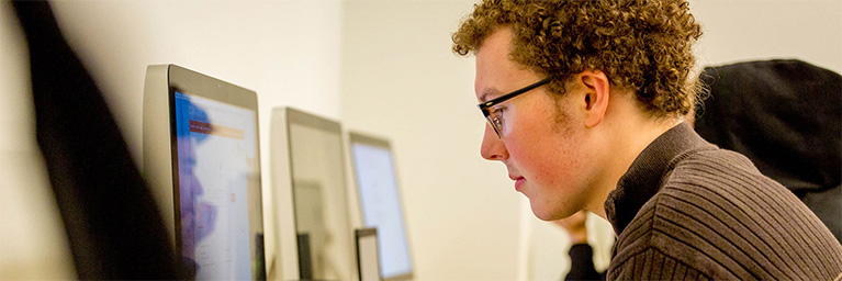 A young man with curly hair and glasses focuses intently on a computer screen at Bethany Lutheran College in Mankato.