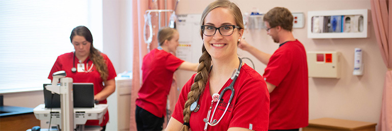 A smiling nurse in red scrubs stands in a medical room on a Minnesota campus, with three colleagues working in the background.