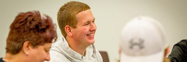 A young man in a gray hoodie smiles while interacting with others in a vibrant Mankato campus community setting.