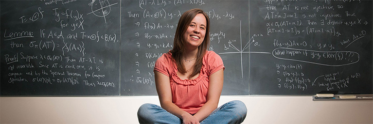 A woman sits cross-legged and smiles in front of a chalkboard covered with math equations at Mankato.