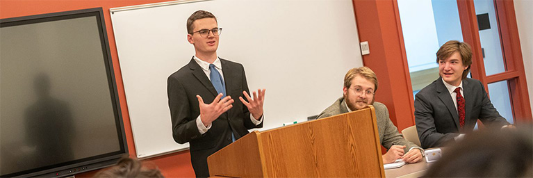 Three young men in suits stand at a podium in a Mankato classroom, presenting to and engaging with the campus community.