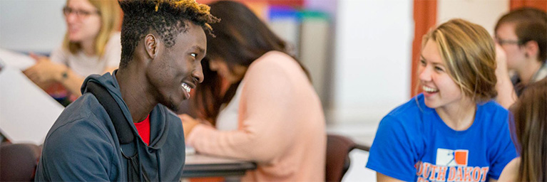 Two students sit facing each other in a Minnesota classroom, smiling and talking, while other students work in the background.