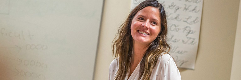 Woman with long brown hair smiling in a classroom, with whiteboards and campus community members engaging in the background.