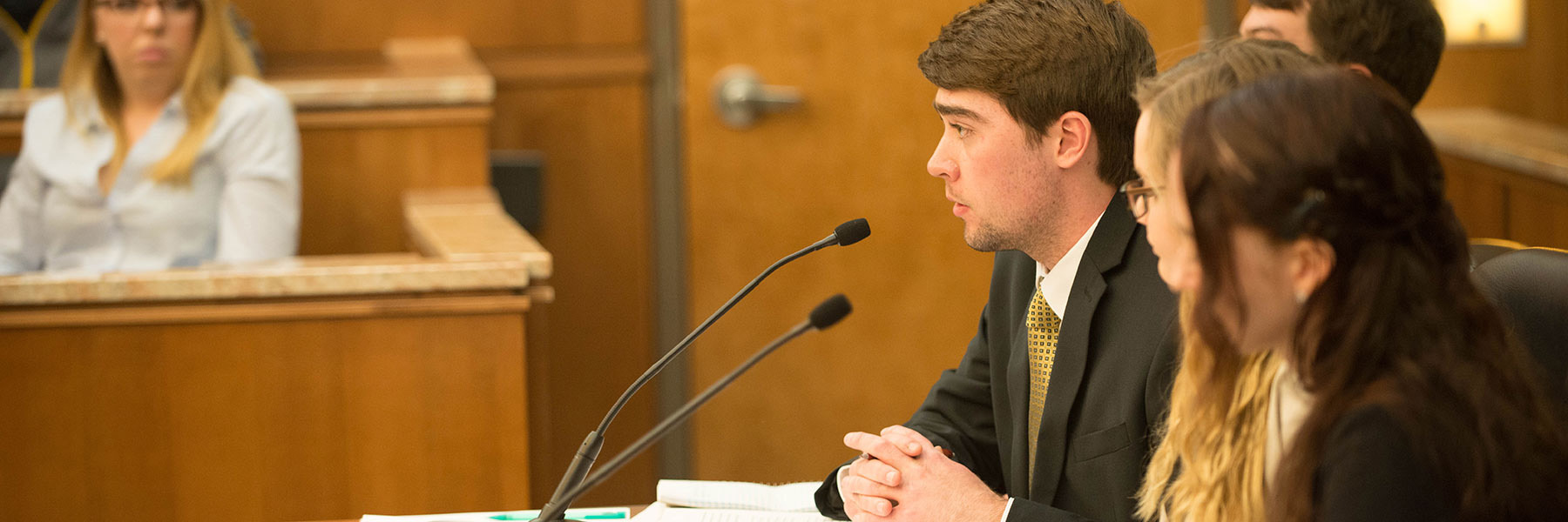 A young man in a suit from Bethany Lutheran College speaks at a courtroom table; a woman sits and listens in the background.