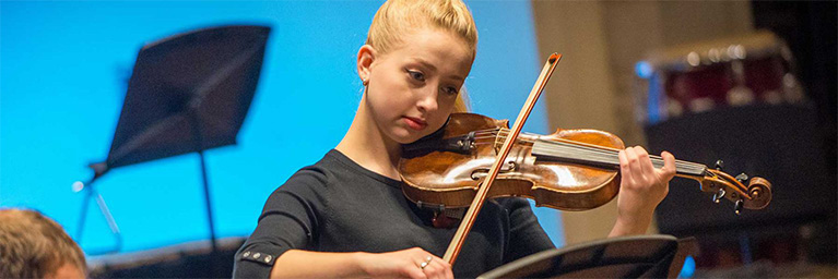 A young woman with blonde hair plays the violin on stage, focusing on sheet music and engaging the campus community.