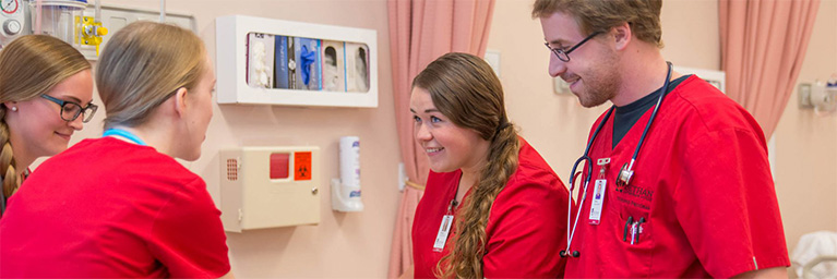 Four medical professionals in red scrubs smile and converse together in a community room at the Mankato campus.