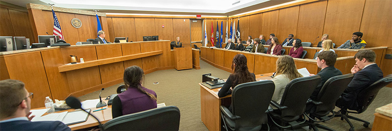 A courtroom scene in Mankato, Minnesota, showing a judge, attorneys, and jury members listening to a speaker at the podium.