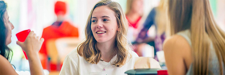 Three young women sit indoors at Bethany Lutheran College, talking and smiling together over food and drinks.