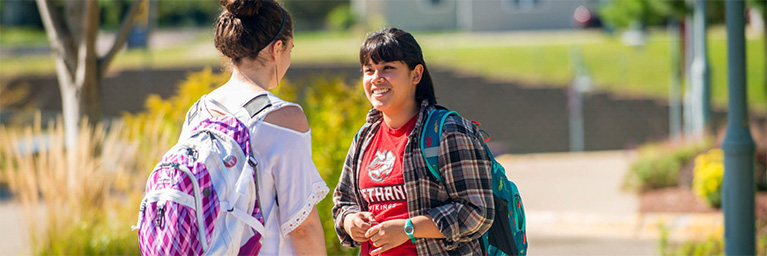 Two students wearing backpacks smile and converse outdoors on a sunny day in Minnesota, demonstrating student engagement.