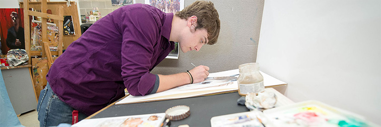 A young man in a purple shirt draws on paper in an art studio at Bethany Lutheran Colleges campus.