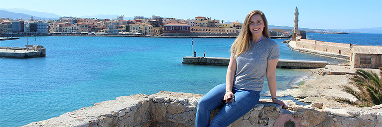 Woman sitting on a stone wall by the sea, with a coastal town and lighthouse in the background, engaging with students.