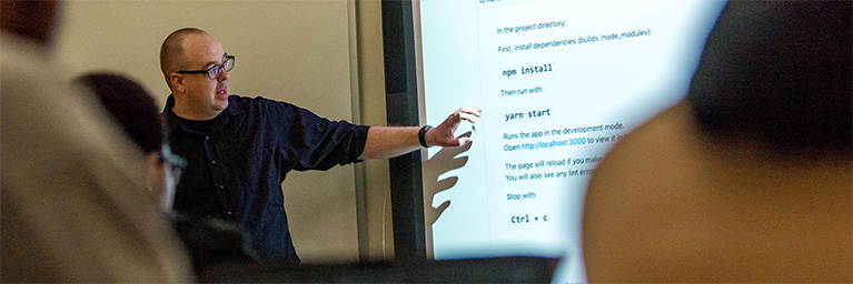A man points to code instructions projected on a screen while teaching a class at Bethany Lutheran College.