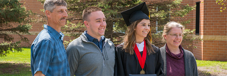 A graduate in cap and gown stands outside with three smiling family members on a sunny day in Mankato.