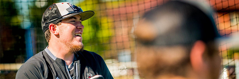 A young man wearing a baseball cap and jersey smiles outdoors near a net, enjoying the campus community in Mankato.
