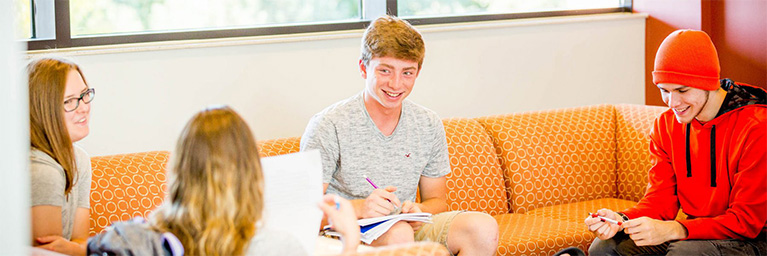 Four students sit on an orange couch at the Mankato campus, smiling and studying together with notebooks.