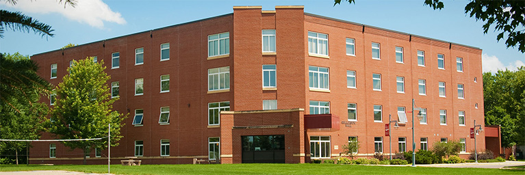 A four-story red brick building at Bethany Lutheran College, featuring numerous windows, surrounded by green grass, trees, and a volleyball net.