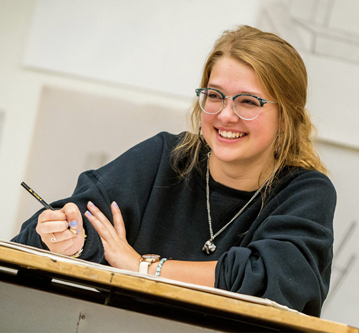 A Bethany Lutheran College student wearing glasses smiles while writing with a pencil at a desk in a classroom.