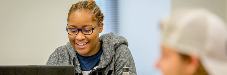 A person wearing glasses and a hoodie smiles while working on a laptop, with the vibrant Mankato campus community in the background.
