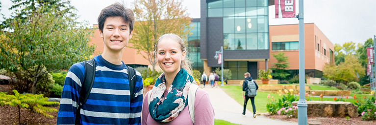 Two smiling students with backpacks stand outside a modern campus building in Minnesota on a cloudy day.