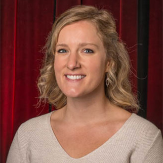 Smiling woman with wavy blonde hair, wearing a beige top, posed in front of red curtains at Bethany Lutheran College in Mankato, Minnesota.
