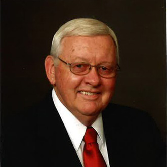 A smiling man with glasses, short gray hair, wearing a dark suit, white shirt, and red tie poses; Bethany Lutheran College in Minnesota.