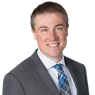 A smiling man in a gray suit and blue plaid tie, photographed against a white background at Bethany Lutheran College.