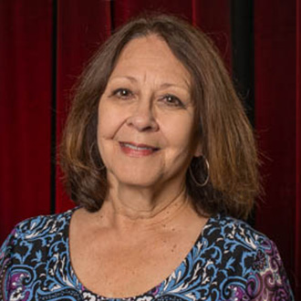 A smiling woman with shoulder-length brown hair, wearing a patterned top, stands in front of red curtains at Bethany Lutheran College.