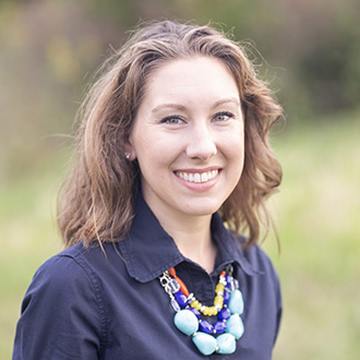 A woman with wavy brown hair, wearing a navy shirt, smiles outdoors on the Bethany Lutheran College campus.