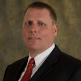 A man wearing a dark suit, white shirt, and red tie stands in front of a blurred campus community background.