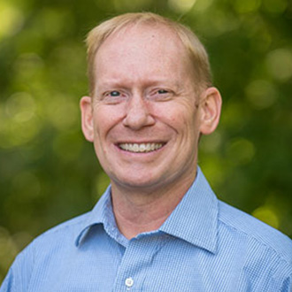 Smiling man with short blond hair wearing a blue collared shirt, standing outdoors at Bethany Lutheran College in Minnesota.