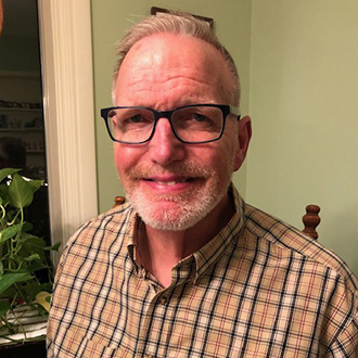 A smiling man wearing glasses and a plaid shirt sits indoors near a window and a plant at Bethany Lutheran College in Mankato.