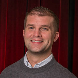 A smiling man wearing a gray sweater over a collared shirt stands in front of a red curtain at Bethany Lutheran College in Mankato.