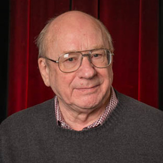 A man wearing glasses and a gray sweater stands in front of a red curtain, smiling slightly in a Mankato campus community setting.