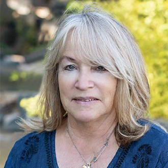 A smiling woman with blonde hair, wearing a blue top, stands outdoors in Mankato, Minnesota. Greenery and sunlight are visible in the background.