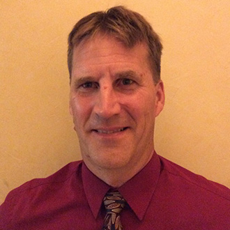 A man with short brown hair, wearing a maroon shirt and tie, smiles in front of a beige background in Mankato, Minnesota.