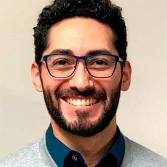 A smiling man with dark curly hair, a beard, and blue glasses, wearing a grey sweater—capturing student engagement at Bethany Lutheran College.
