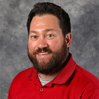 A man with dark hair and a beard, wearing a red collared shirt, smiles in front of a gray background at Bethany Lutheran College.